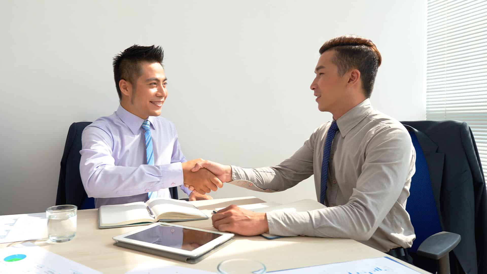 Dua pria profesional berkemeja dan dasi sedang berjabat tangan sambil tersenyum di meja kerja ruang kantor.