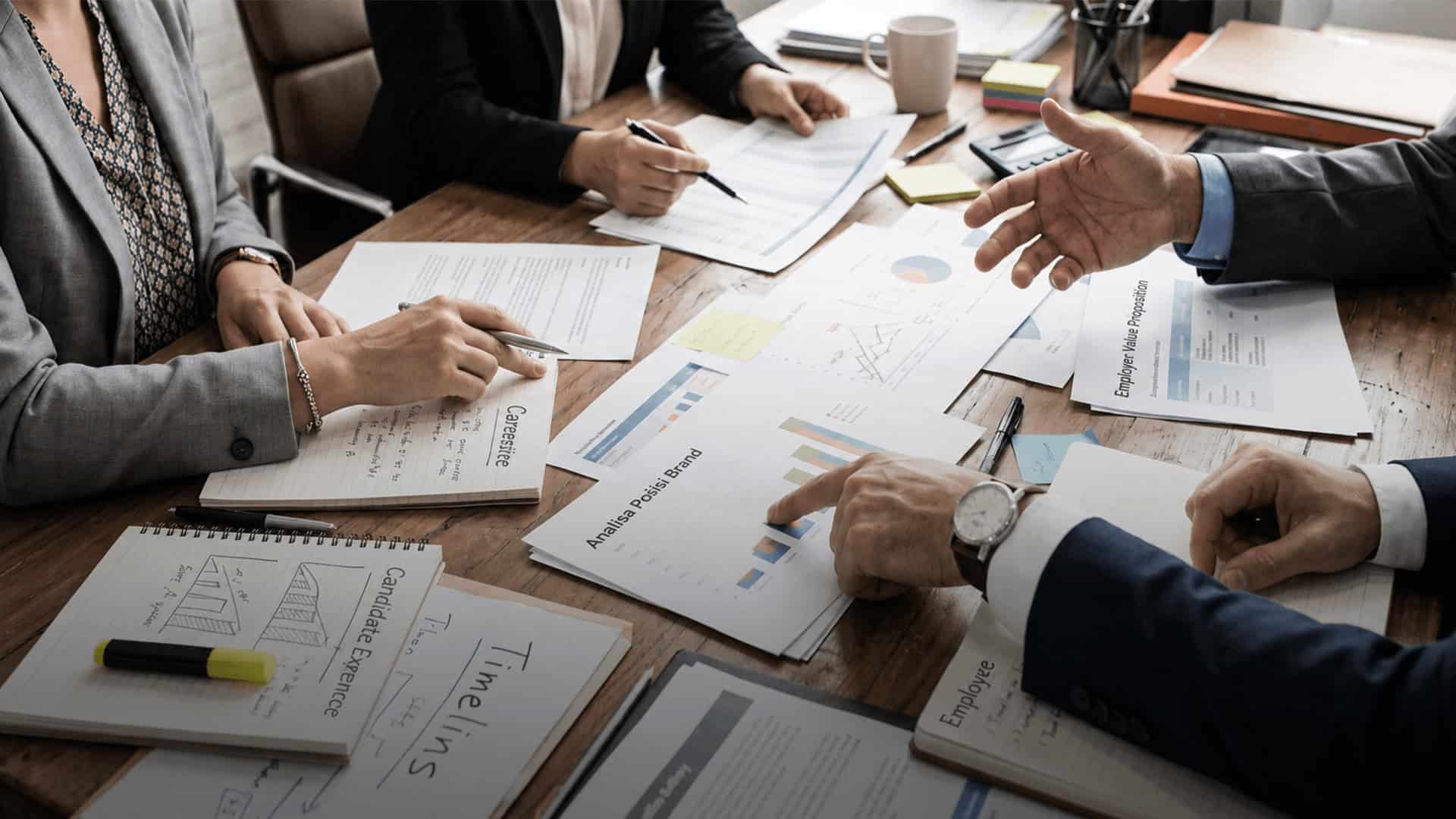 Tim profesional sedang berdiskusi sambil menunjuk dokumen dan grafik analisis merek di meja rapat.
