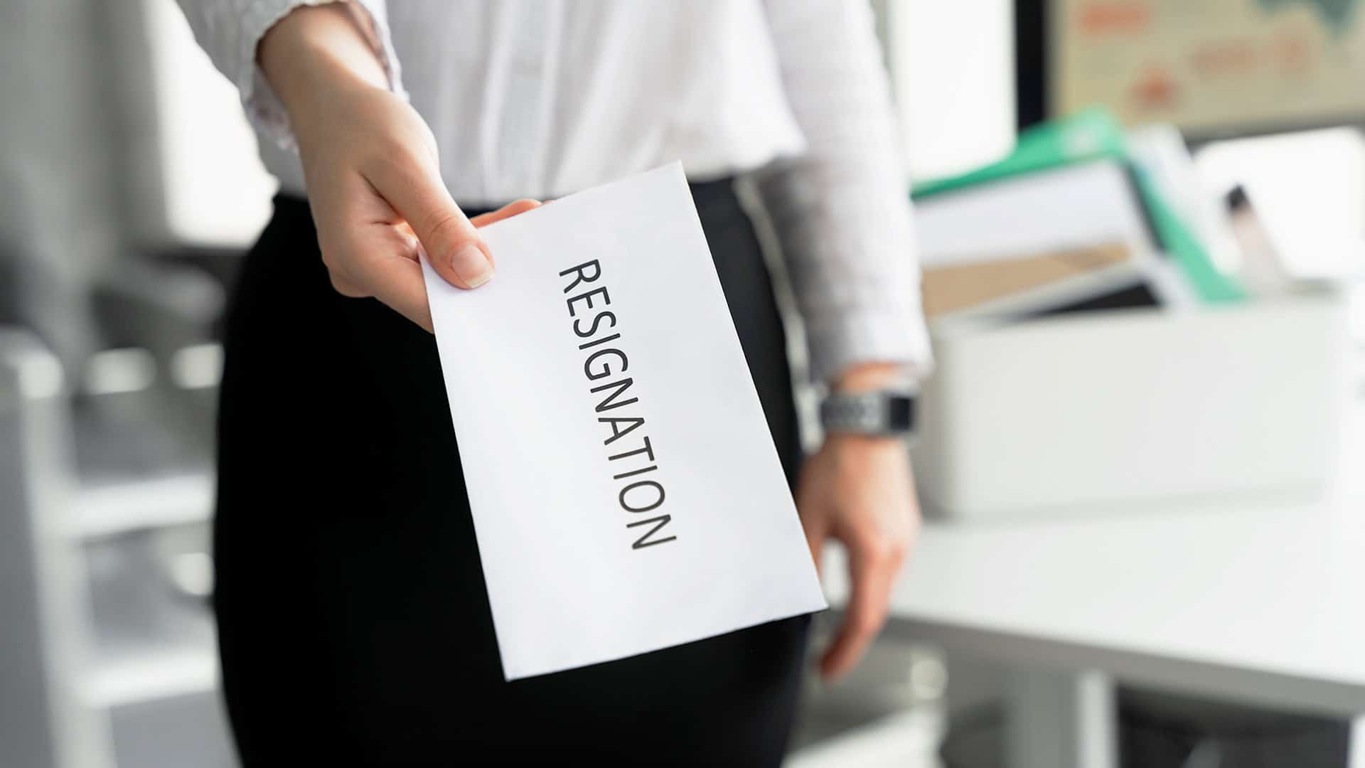 Seorang pekerja menyerahkan amplop putih bertuliskan RESIGNATION di area kantor.